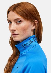 Blå atletisk jakke med høj krave, featuring logoet "Peak Performance" i sort på kraven. Glat tekstur, tætsiddende design.