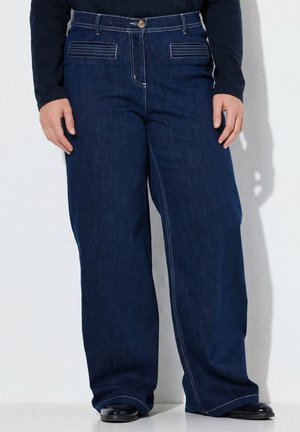 Person wearing wide-leg dark blue jeans with white stitching, standing against a white wall, hands relaxed by sides.