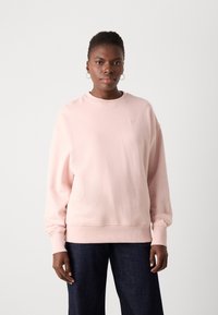 Lys rosa genser med ribbestrikkede ermer og nederkant, med et enkelt brodert logo på brystet. Myk stoffkvalitet, løs passform.