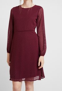 Bordeauxfarbenes Kleid mit langen durchsichtigen Ärmeln, strukturiertem Fischgrätmuster, runder Halsausschnitt und einer taillierten Akzentuierung, knielang.