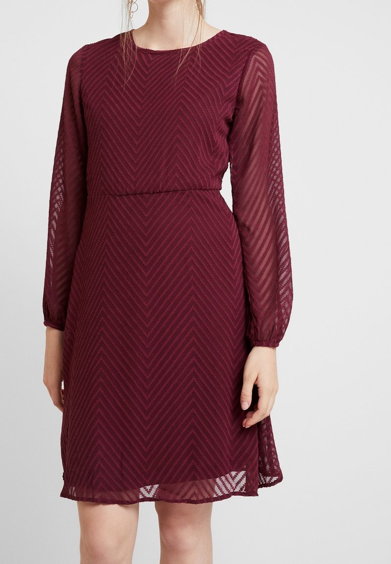 Bordeauxfarbenes Kleid mit langen durchsichtigen Ärmeln, strukturiertem Fischgrätmuster, runder Halsausschnitt und einer taillierten Akzentuierung, knielang.