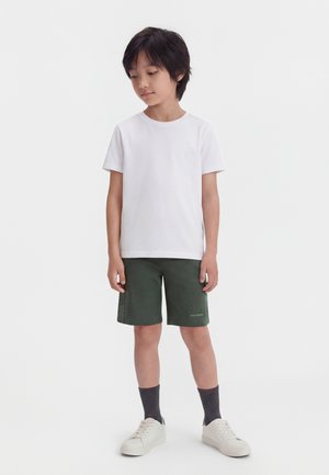 Mladý chlapec stojí, má na sebe obyčajné biele tričko, tmavé zelené šortky, tmavé ponožky a biele tenisky na bielom pozadí.