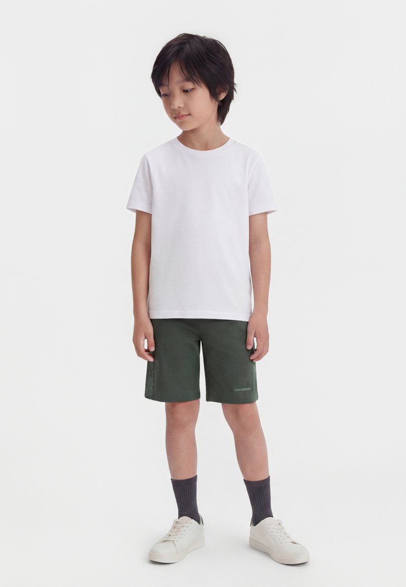 Νέος αγόρι στέκεται, φοράει απλό λευκό μπλουζάκι, σκούρα πράσινα σορτς, σκουρόχρωμες κάλτσες και άσπρα αθλητικά παπούτσια σε λευκό φόντο.