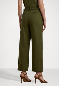 Calças largas em tecido verde-azeitona, com cintura alta, bolsos traseiros com botões e uma bainha limpa. Combinadas com sandálias de salto castanhas.
