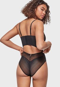 Crni bodysuit s prilagođenim dizajnom, s mrežastim preklopom s dijamantnim uzorcima. Vidljive su podesive naramenice i detalj s niskim leđima.