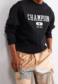 Black sweatshirt with white "CHAMPION" text, round logo, and ribbed cuffs, paired with beige, patterned cargo pants featuring multicolored accents.