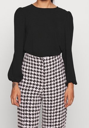 Mujer con blusa negra de manga larga con hombros abullonados y pantalones con estampado de pata de gallo en blanco y negro.