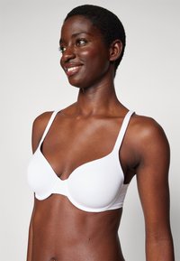 Weißer nahtloser BH mit verstellbaren Trägern, weichem Material und niedrigem Ausschnitt. Verfügt über geformte Cups und ein minimalistisches Design.