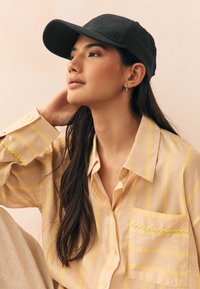 Sort baseballkasket, lysegul skjorte med striber og en lomme med limegrøn kant, glat stof, model i profil mod en blød baggrund.