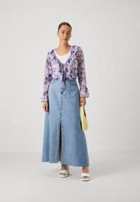 Purple floral sheer blouse with ruffled neckline, paired with a high-waisted blue denim maxi skirt featuring a front slit. White platform sandals.