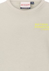 Jasno beżowa bluza dresowa wykonana z miękkiego materiału. Posiada okrągły dekolt oraz tekst "RETOUR ESSENTIAL IT STARTS" w fluorescencyjnym żółtym na piersi.