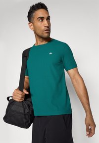 Türkises kurzärmliges Shirt mit rundem Halsausschnitt und dezentem Logo; das Modell hält eine schwarze Tasche; der Stoff wirkt glatt und eng anliegend.