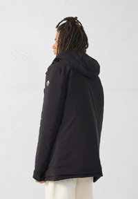 Persoon met dreadlocks die een zwarte hoodie en crèmekleurige broek draagt, naar links kijkt tegen een effen witte muur als achtergrond.