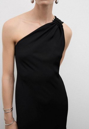 Femme portant une robe noire à une épaule avec des ornements en argent en forme de billes et de grandes boucles d'oreilles en argent, sur un fond blanc uni.