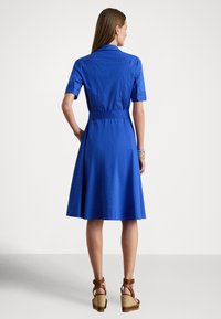 Mėlyna medvilninė marškinių tipo suknelė trumpomis rankovėmis, su diržu ties liemeniu ir išleistu sijonu. Dėvima su rudos spalvos aukštakulniais sandalais. Peržiūra iš nugaros, lygi tekstūra.