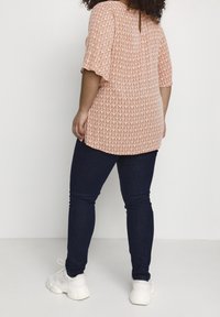 Woman with curly hair wearing an orange patterned short-sleeve blouse, dark blue fitted jeans, and white sneakers standing facing away.