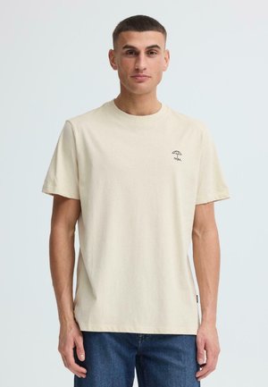 Jonge man draagt een lichtbeige T-shirt met een klein paraplu-ontwerp en de tekst "relax", gecombineerd met een blauwe spijkerbroek, staand tegen een effen achtergrond.