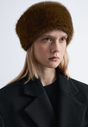 Brown fur hat with a plush texture and rounded shape, worn by a model in a black coat against a plain background.