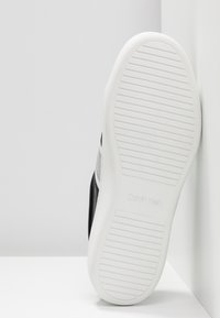 Semelle en caoutchouc blanc d'une basket Calvin Klein avec des rainures horizontales et le nom de la marque embossé au centre, sur un fond blanc en angle.