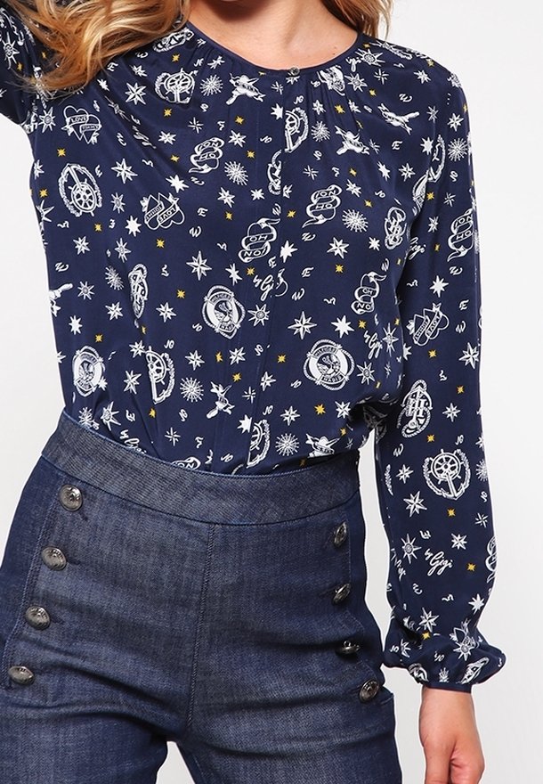 Femme portant un chemisier bleu marine avec des imprimés nautiques blancs et un pantalon bleu foncé taille haute avec des rangées doubles de boutons à l'avant.