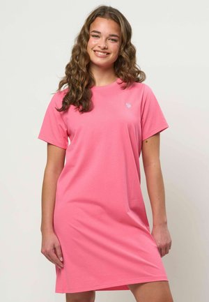 Nuori nainen, jolla on pitkät laineikkaat hiukset ja joka hymyilee. Hänellä on päällään vaaleanpunainen, lyhythihainen ja rento mekko, ja taustalla on yksivärinen vaalea pinta.