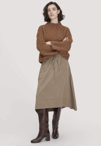 Frau mit kurzem, lockigem Haar trägt einen braunen Strickpullover, einen beigen Midirock und dunkelbraune knielange Stiefel, steht mit verschränkten Armen.