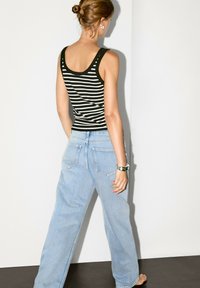 Een vrouw, gezien van achteren, in een gestreepte tanktop en lichtblauwe wijde jeans, staat tegen een effen achtergrond.