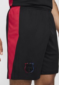 Sorte atletiske shorts med et rødt sidelogo, mesh tekstur, elastisk talje og et stilrent sort og farvet våbenskjoldlogo i kanten.