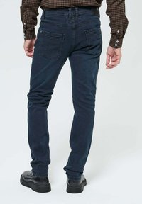 Homme portant un jean bleu foncé, des chaussures noires et une chemise à carreaux marron, debout de dos sur un fond clair uniforme.