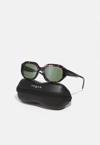 Ochelari de soare octogonali cu rame negre, cu model de țestoasă închisă și lentile verzi, așezați pe un etui rigid negru elegant, cu închidere cu fermoar.
