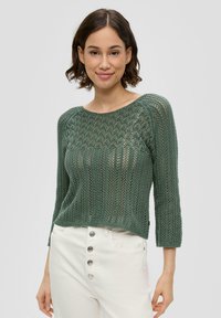 Grüner Strickpullover mit strukturiertem Muster, Dreiviertelärmel und Rundhalsausschnitt. Kombiniert mit weißen, hochsitztenden Hosen mit Knopfakzenten.