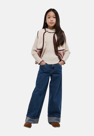 Jeune fille debout, les mains dans les poches, portant un pull crème à bords volantés, un gilet patchwork, un jean large avec revers et des chaussures marron.