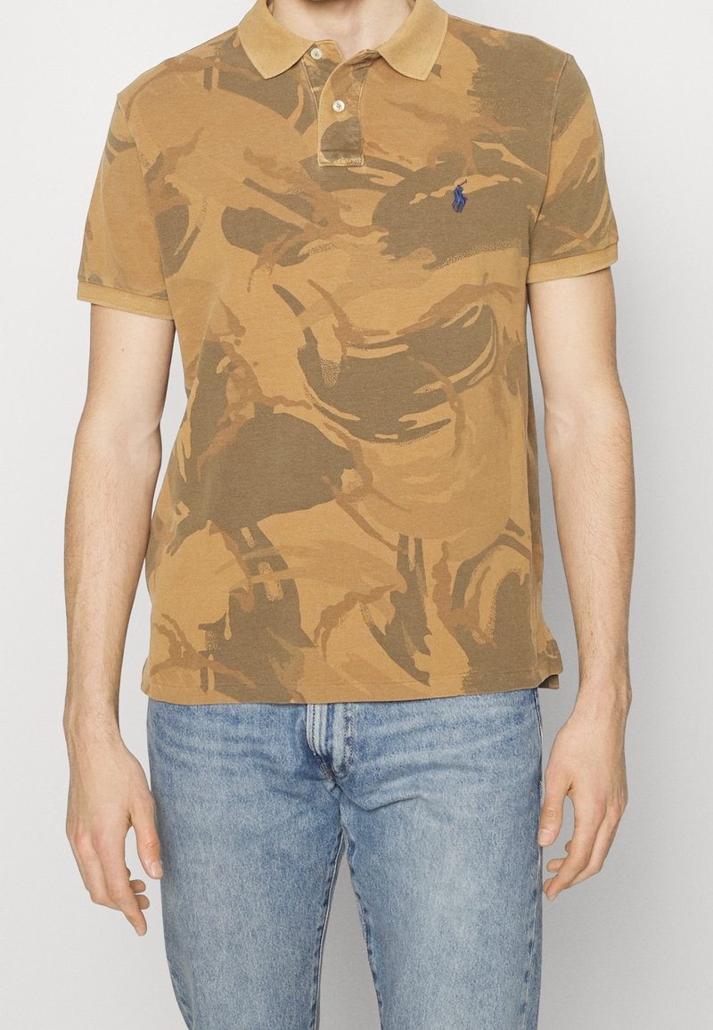 Man med en brun kamouflage-mönstrad pikétröja med en blå broderad logotyp, i kombination med ljusblå denimjeans.