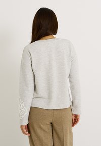 Sudadera de color gris claro con un ajuste holgado, cuello acanalado y bordado de flores en la manga izquierda. Textura suave, combinada con pantalones beige.