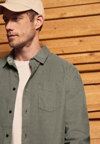 Camisa de color verde claro, abotonada, con un bolsillo en el pecho, hecha de una tela texturizada. Lleva puesta sobre una camisa de color claro, con paneles de madera de fondo.