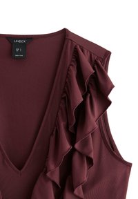 Burgundy ärmlös topp med djup V-ringning och volangdetalj längs vänster sida, tillverkad av smidigt tyg. Varumärkesetikett synlig.