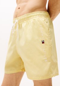 Żółte szorty kąpielowe w paski z elastycznym pasem, sznurkiem do regulacji i białym haftowanym logo "Tommy Hilfiger" z boku.