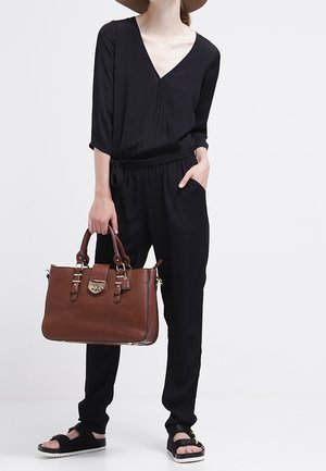 Svart långärmad jumpsuit med V-ringning, avslappnad passform och draperad midja. Brun läderhandväska med guld detaljer och överhandtag. Svarta sandaler.