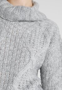 Hellgrauer, gestrickter Pullover mit einem gerippten Kragen, der ein strukturiertes Muster und einen Zopfstrickbereich auf der Vorderseite aufweist.