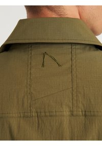 Zbliżenie na tylną część kołnierza oliwkowej zielonej kurtki z naszytym logo i widocznymi detalami szwów.