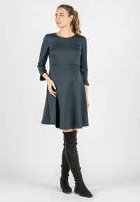 Dunkelgraues knielanges Kleid aus glattem Stoff mit Dreiviertelärmeln und ausgestellt geschnittenem Saum. Kombiniert mit schwarzen Overknee-Stiefeln.