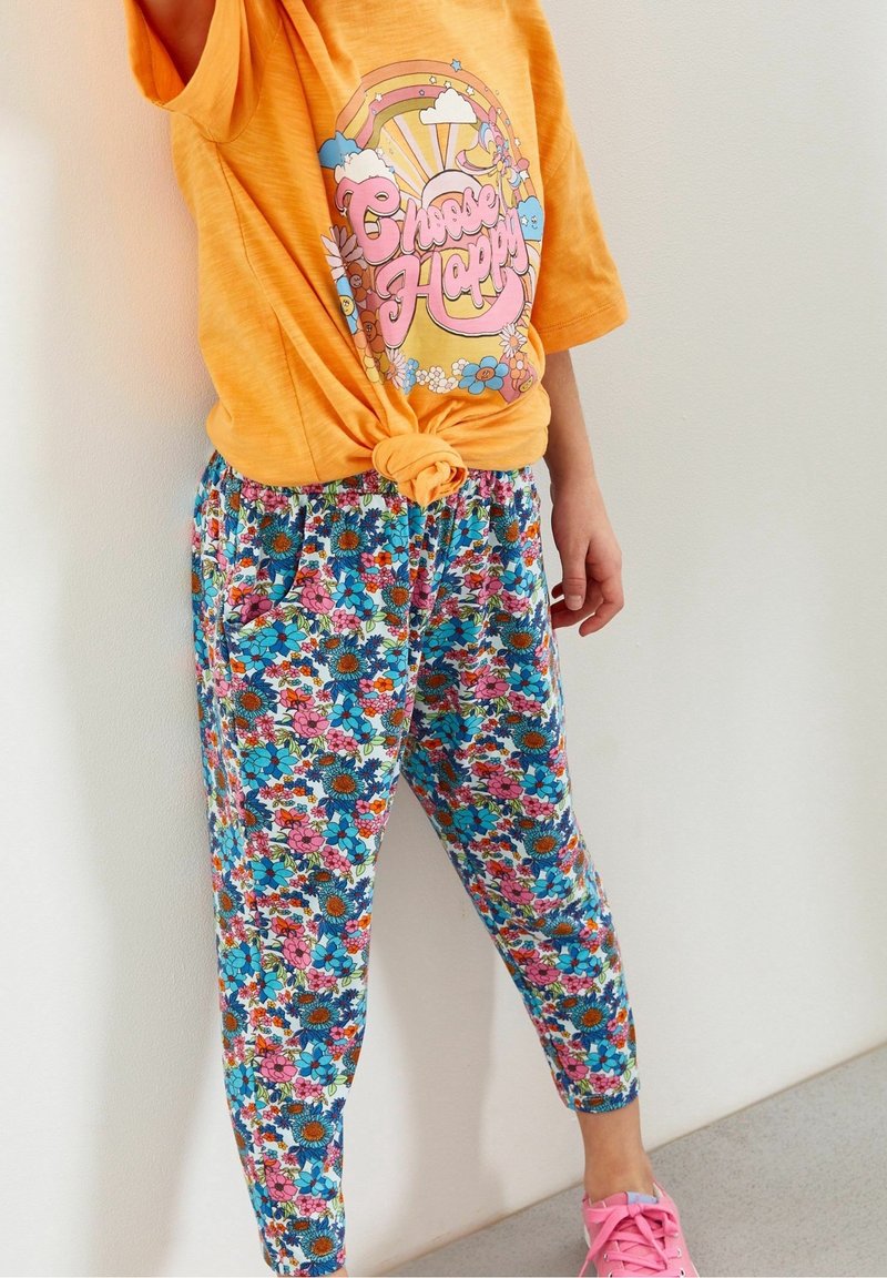 Chemise orange vif avec le texte « Choose Happy » et un graphisme arc-en-ciel, associée à un pantalon floral coloré et des chaussures roses, portée par une personne debout.