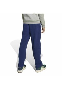 Eine Person trägt marineblaue Jogginghosen, einen grauen Sweatshirt und grüne Adidas-Sneaker, steht mit dem Rücken zur Kamera vor einem weißen Hintergrund.