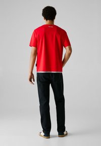 Vīrietis stāv ar muguru pret skatītāju, ģērbies sarkanā Pepe Jeans T-kreklā, melnās džinsās un baltos sporta apavos uz vienkārša pelēka fona.