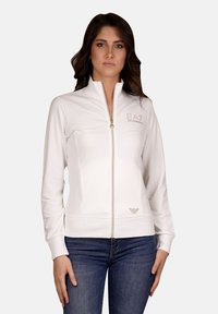 Giacca bianca con zip e colletto alto, dal design aderente. Presenta un logo in strass e polsini a costine. Tessuto dalla texture liscia.