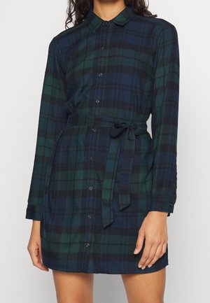 Kobieta ubrana w sukienkę-koszulę w kratę w kolorach ciemnej zieleni i granatu, z długim rękawem, zapinaną na guziki z przodu i z paskiem wiązanym w talii.