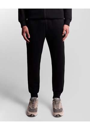 Personne portant un pantalon de jogging noir et des baskets New Balance beiges, debout sur un fond gris clair uni.