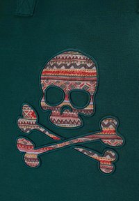Tela de color verde oscuro con un diseño destacado de calavera y huesos cruzados. La calavera está decorada con patrones geométricos coloridos, aportando detalles.