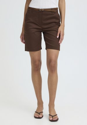 Vrouw die bruine knielange shorts draagt met een smalle riem, een witte mouwloze top en bruine teenslippers, op een witte achtergrond.