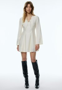Cremefarbenes Minikleid mit schnürbarem Ausschnitt und Glockenärmeln. Kombiniert mit knielangen schwarzen Lederstiefeln. Glatte Textur, strukturierte Form.